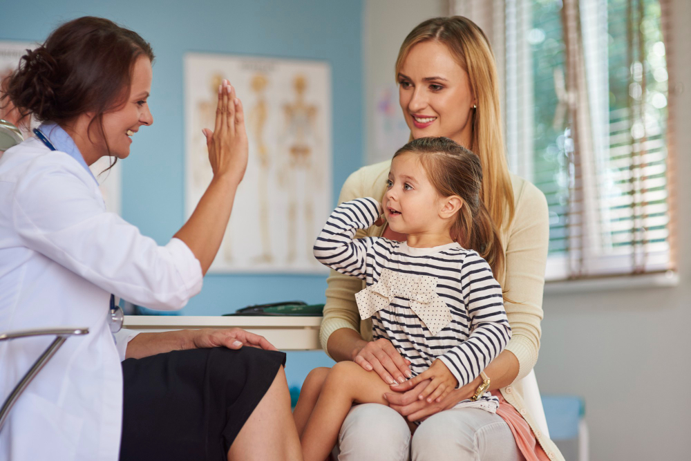 Una doctora sonriente choca la mano con una niña sentada en las piernas de su mamá