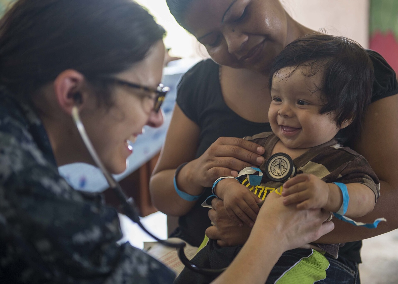 Una proveedora escucha el corazón de un bebé sonriente mientras su madre lo sostiene